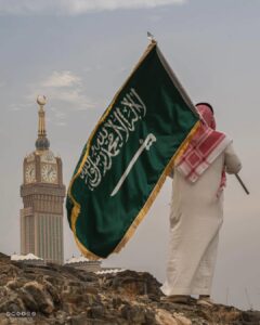 A Saudi Man Standing atop a hill, proudly hoisting the Saudi national flag with the iconic Makkah Clock Tower in the background - Image via Social media